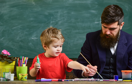 Teacher with beard, father teaches little son to draw in classroom, chalkboard on background. Art lesson concept. Talented artist spend time with son. Child and teacher on busy face painting, drawingの写真素材