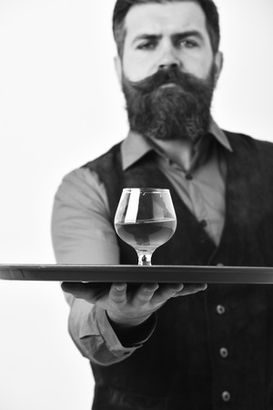Barman in vintage vest with serious face serves cognac. Man with beard holds glass with alcohol on white background.の写真素材