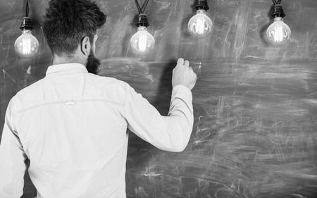 Guy busy with writing on chalkboard surface. Announcement concept. Man with beard stand in front of chalkboard, rear view. Bearded hipster in shirt writing on chalkboard, copy space. Guy busy with writing on chalkboard surfaceの写真素材