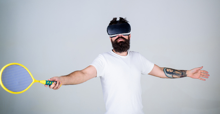 Guy with VR glasses play tennis with racket and ball. Man with beard in VR glasses beating pitch, grey background. Hipster on busy face use modern technology for sport games. Virtual sport conceptの写真素材