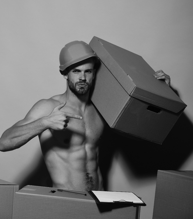 Man with beard stands naked on pink background. Macho with cool face wears helmet and has folder. Guy with sexy torso holds box on shoulder and points to it. Sexuality and moving concept.の写真素材