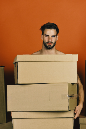 Delivery and moving in concept. Man with strong arms holding cardboard boxes. Macho with beard and serious face. Guy with pile of cartons isolated on red background.の写真素材