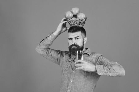 Guy presents homegrown harvest. Gardening and fall crops concept. Man with beard holds bowl of fruit on head and juice on blue background. Farmer with serious face holds fresh apple juice and apples.の写真素材
