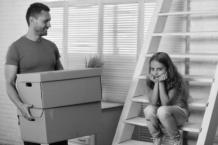 Kid and guy move in or move out. New home and family concept. Daughter and father hold boxes and unpack or pack. Girl and man with smiling and grumpy faces in room with ladder and window on background.の写真素材