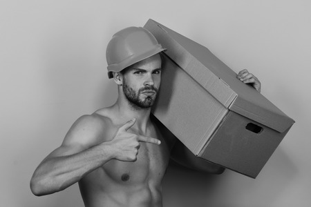 Sexuality and moving concept. Man with beard stands on pink background, points to box. Guy with sexy naked torso lifts boxes. Macho with selfsatisfied face wears helmet and has folder.の写真素材