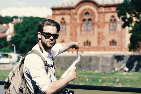Travelling and landmark concept. Man with serious face holds map and camera. Tourist does sightseeing looking for tourists attractions. Traveller points at historical building on background, defocused.の写真素材