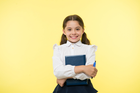 I am ready for school. Child school uniform smart kid done with homework. Girl cute happy face yellow background. Child girl school uniform clothes holds book and pen. Back to school conceptの写真素材
