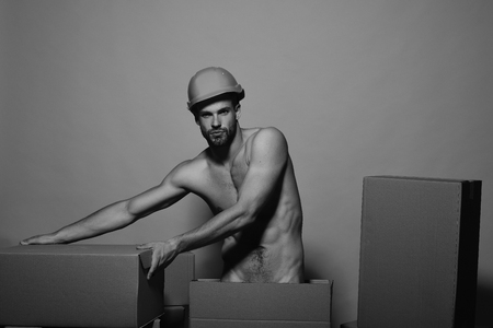 Guy with sexy naked torso moves boxes. Macho with serious face wears helmet. Man with beard stands on yellow background. Sexuality and building concept.の写真素材