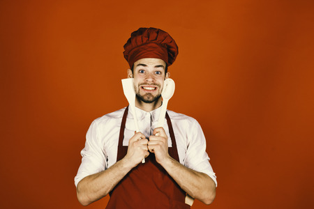 Kitchenware and cooking concept. Chef with excited face holds wooden spoon and spatula near face on red background. Man in cook hat and apron holds cooking tools. Cook works in kitchen.の写真素材