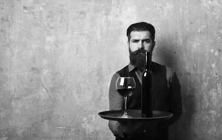 Waiter with glass and bottle of red wine on tray.. Man with beard holds wine on beige wall background, copy space. Barman with serious face serves wine glass. Service and restaurant drinks concept.の写真素材