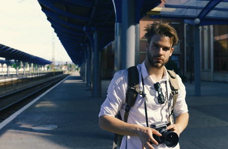 Man with beard holds photocamera on station background. Urban photo and travelling concept. Tourist ready to take picture of cityscape. Young traveller or photographer waits for train.の写真素材
