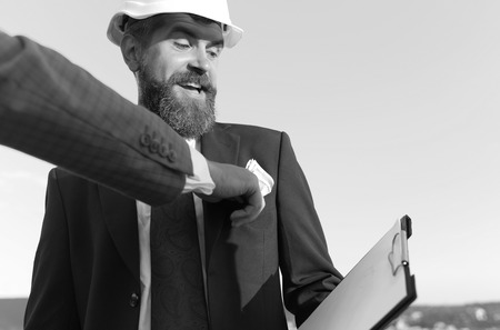 Architect in suit holds clip folder on blue sky background. Construction and bribery concept. Male hand puts money into coworkers pocket. Engineer with beard and white helmet smiles and looks at cash.の写真素材