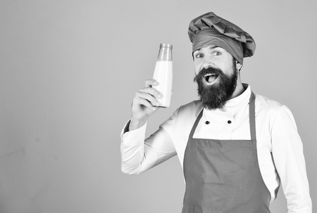 Nutritious lactose concept. Cook with excited face in burgundy hat and apron holds milk. Chef with milkshake or yoghurt. Man with beard holds glass bottle on red background, copy space.の写真素材