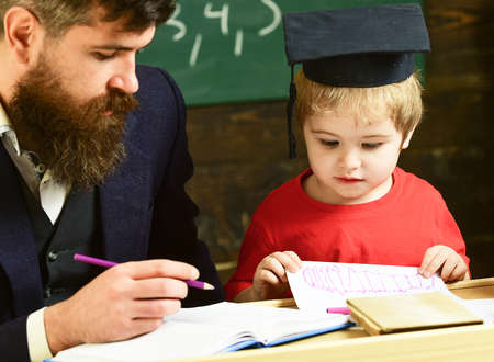 Teacher in formal wear and pupil in mortarboard in classroom, chalkboard on background. Enthusiastic kid studying with teacher. Father checking homework, helps to boy, son. Homeschooling concept.の写真素材