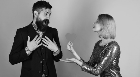 Girl and bearded man with surprised face share paper heart card for valentines date. Love and holidays concept. Girlfriend offers her love to boyfriend. Couple in love holds heart on blue background.の写真素材