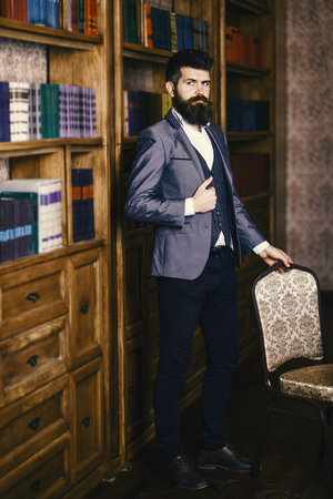 Businessman sits in vintage interior, holds book and cup of tea. Bearded man in luxury suit in his cabinet. Mature man with calm face enjoys reading. Luxury lifestyle, vintage fashion, tea concept.の写真素材
