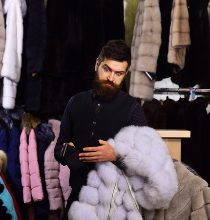 Man with beard and mustache in fur shop. Guy with shocked face looks into empty wallet and holds mink fur coat. Macho with wallet in front of luxury furry clothes. Luxury clothing concept.の写真素材