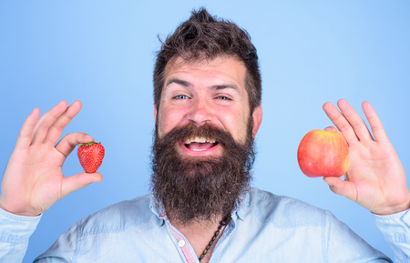 Man bearded smiling holds apple and strawberry in hands blue background. Vitamin fruit nutrition concept. Healthcare dieting vitamin. Fruit and berry in hands healthy alternative. Summertime benefits.の写真素材