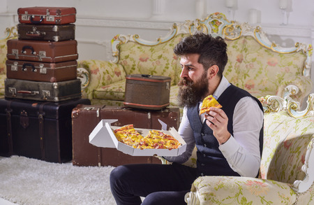 Bon appetit concept. Man with beard and mustache holds box with tasty fresh hot pizza. Macho in classic clothes hungry, holds slice of cheese pizza, eats, enjoying taste, interior background.の写真素材