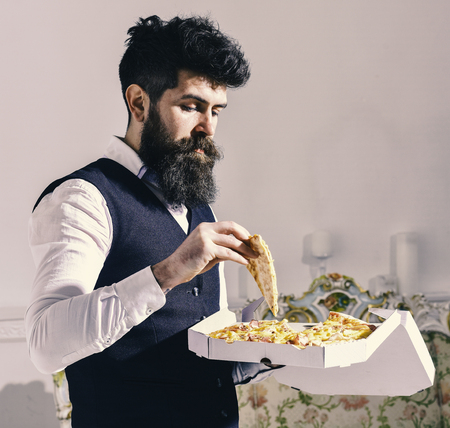 Food delivery concept. Macho in classic clothes hungry, holds slice of cheese pizza, eats, enjoying taste, interior background. Man with beard and mustache holds box with tasty fresh hot pizza.の写真素材