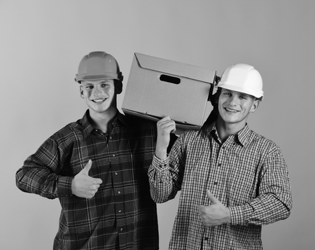 Twin brothers in helmets show thumbs up. Delivery, warehouse and packaging concept. Siblings with carton box work as deliverymen. Men with happy faces carry cardboard box on pink background.の写真素材