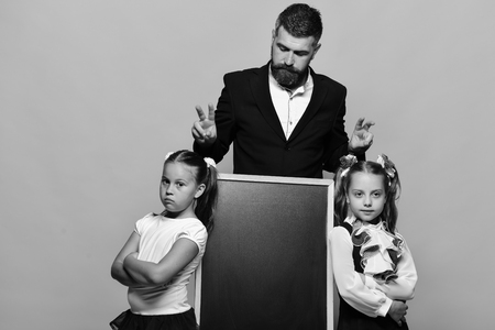 Schoolgirls near blackboard and guy on yellow background. Back to school concept. Girls and man with grumpy and serious faces. Teacher with beard and kids with ponytails next to blackboard, copy space.の写真素材