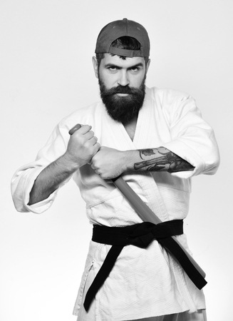 Baseball player with serious face puts green baseball bat behind black belt. Master gets ready to fight. Martial arts concept. Man with beard in white kimono and green cap on white background.の写真素材