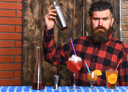 Man in checkered shirt on wooden texture background. Barman and cocktails concept. Barman with long beard and mustache and stylish hair with strict face holding shaker and make alcoholic cocktail.の写真素材