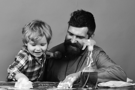 Cleaning concept. Guy with beard and kid in checkered shirt at table near cleaning spray. Man with smiling child on green background. Son with father cleaning together with sponges.の写真素材