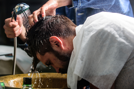 Man bearded client of hipster barbershop. Barbershop concept. Barbers hands washing hair of bearded hipster. Man with beard and mustache with towel on shoulders, male hands with shower on background.の写真素材