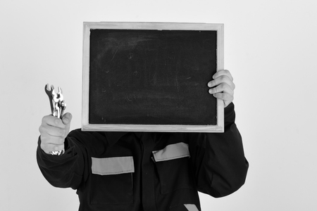 Man hides face behind empty chalkboard on white background. Plumber with metallic spanner equipment in hand. Mechanic or repairer holds blackboard with copy space. Occupation and advertising concept.の写真素材