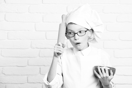 young boy small cute cook chef in white uniform and hat on surprised face with glasses holding red bowl with rolling pin on brick wall background, copy spaceの写真素材