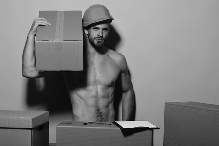 Man with beard stands on pink background, copy space. Guy with sexy naked torso holds box on shoulder. Seduction and moving concept. Macho with interested face wears helmet and has folder.の写真素材