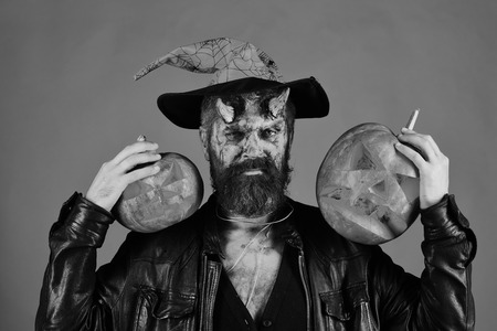 Demon with horns and strict face holds jack o lanterns. Man wearing scary makeup and witchers hat...