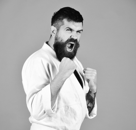 Young sporty man in white kimono isolated on red background. Athlete with beard and raging face holds fists in defence. Japanese sports and martial arts conceptの素材 [FY310104379408]