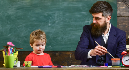 Child and teacher on busy face painting, drawing. Art lesson concept. Teacher with beard, father teaches little son to draw in classroom, chalkboard on background. Talented artist spend time with son.の写真素材