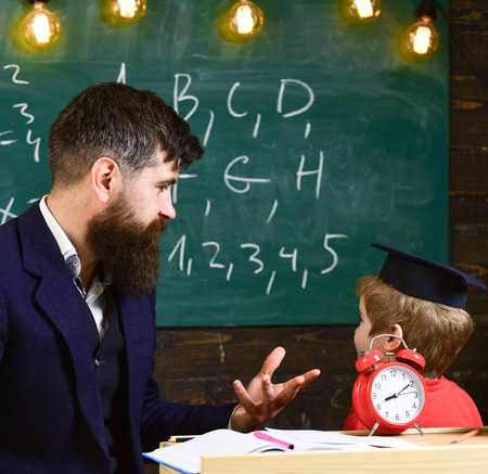 Individual lesson concept. Teacher with beard, father teaches little son in classroom, chalkboard on background. Boy, child in graduate cap looks at scribbles on chalkboard while teacher explains.の写真素材