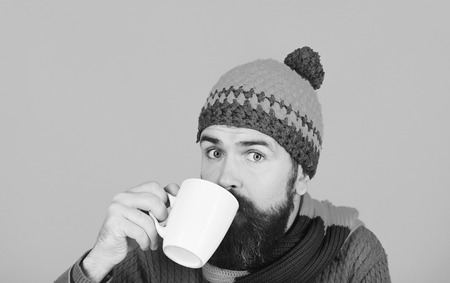 Autumn coffee and home barista concept. Caffeinated or decaf beverages idea. Man in warm hat holds white cup on orange background, copy space. Hipster with beard and curious face has fragrant coffee.の写真素材