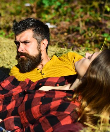 Girl and bearded guy or happy lovers lie on dry grass. Couple in love spends time in park. Man and woman with serious faces on nature background. Autumn outdoor leisure and love conceptの写真素材