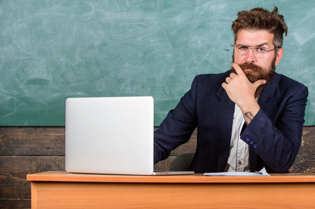 I am your new teacher. Teacher bearded hipster with eyeglasses sit in classroom chalkboard background. School teacher ready to start lesson. Teacher sit at desk with laptop. Back to school concept.の写真素材