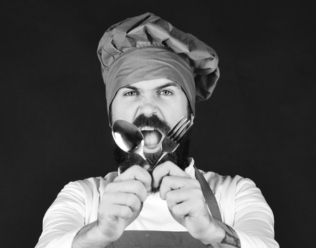 Kitchen utensils concept. Man with beard holds kitchenware on black background. Chef holds cutlery crossed. Cook with angry face in burgundy hat and apron holds spoon and fork.の写真素材