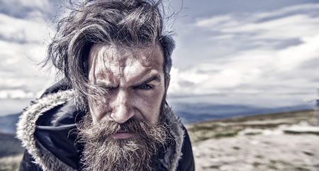 Handsome man hipster or guy with beard and moustache on serious face in hat and jacket outdoor on mountain top against cloudy sky on natural background, copy spaceの写真素材