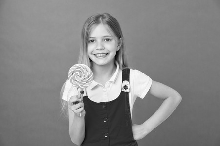 Girl eating big candy on stick or lollipop. Sweet childhood concept. Girl on smiling face holds giant colorful lollipop in hand, violet background. Kid with long hair likes sweets and treats.の写真素材