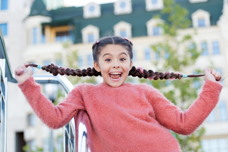 Funny cute girl in city showing her long beautiful braids. Playing girl with very happy smile on her face. Having some fun time outside. Photo in downtown. Happy childhood.の写真素材