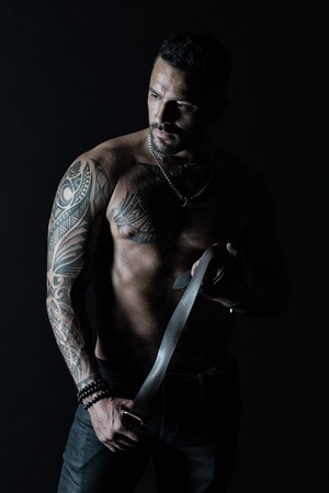 Fashion model with leather belt in jeans. Man with tattoo design on skin. Bearded man with muscular torso. Sportsman with tattooed arm and chest. Bodycare with fitness and sport in vintage filter.の写真素材