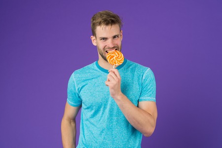 Man bearded adult likes lollipop. Sweet tooth concept. Sugar harmful for health. Guy hold lollipop candy violet background. Man handsome macho eat big colorful sweet lollipop. Taste of childhood.の写真素材