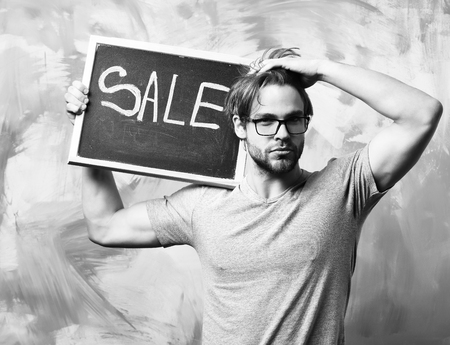 Bearded man, short beard. Caucasian sexy serious macho man with moustache in glasses wearing gray shirt holding black board with sale inscription on colorful studio backgroundの写真素材