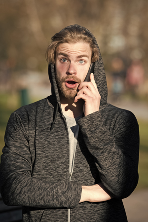 Guy with surprised face talk on smartphone in park, communication. Bachelor in hood with mobile phone on sunny day, fashion. Modern life, new technology, communication. Fashion, style, trendの写真素材