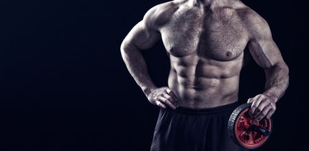 bodybuilder man or sexy muscular guy with athlete body, torso and chest training in gym, holds red six pack trainer, roller, wheel trimmer, sports equipment with hilt and rubber on black backgroundの写真素材
