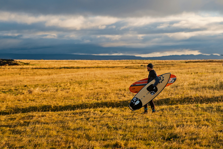 Reykjavik, Iceland - October 14, 2017: Man surfer walk on grass field in reykjavik, iceland. Man with surfboards on autumn landscape. Sportsman on cloudy grey sky. Sport activity and energy. Vacation and wanderlust.の写真素材